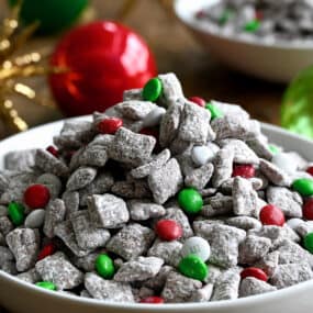 Christmas Puppy Chow with red, green and white M&Ms in a white bowl next to holiday ornaments