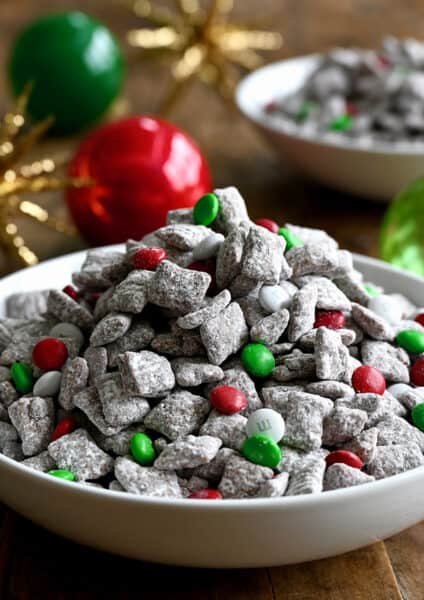 Christmas Puppy Chow with red, green and white M&Ms in a white bowl next to holiday ornaments