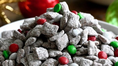 Christmas Puppy Chow with red, green and white M&Ms in a white bowl next to holiday ornaments