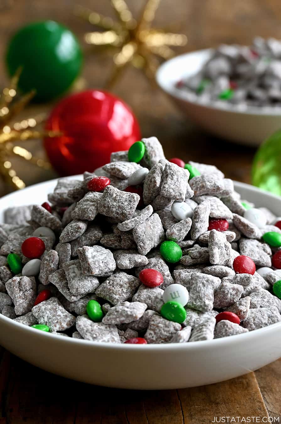 Christmas Puppy Chow with red, green and white M&Ms in a white bowl next to holiday ornaments