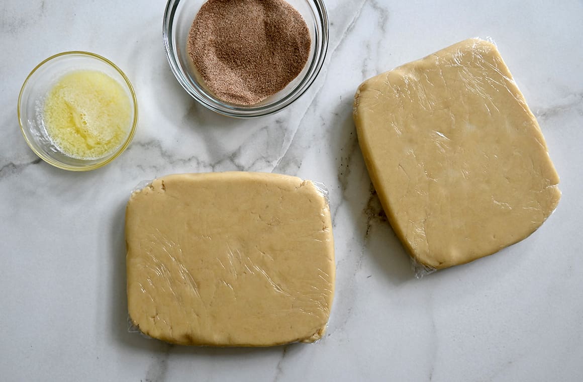 A top-down view of two chilled dough squares wrapped in plastic wrap next to a bowl containing melted butter and a bowl containing cinnamon-sugar mixture