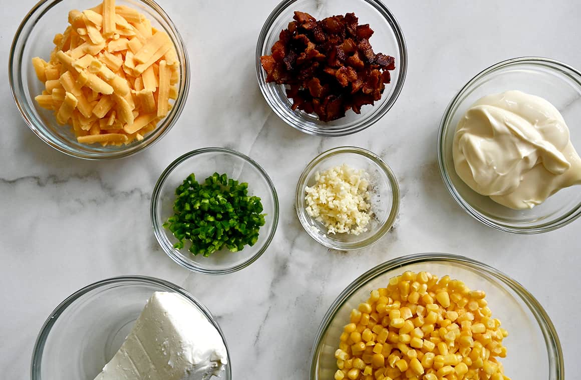 A top-down view of various sizes of bowls containing shredded cheddar cheese, bacon pieces, mayo, corn kernels, minced garlic, chopped jalapeño and a block of cream cheese