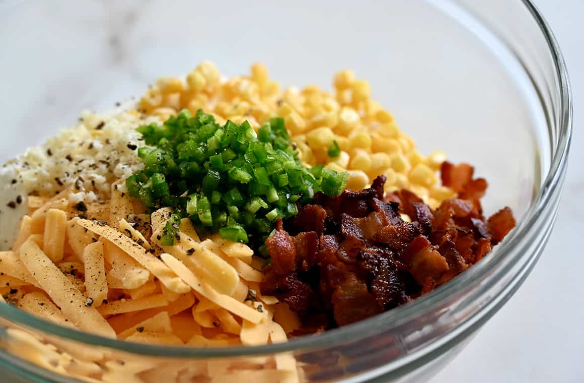 A clear bowl containing yellow corn kernels, chopped jalapeño, bacon pieces and shredded cheddar cheese