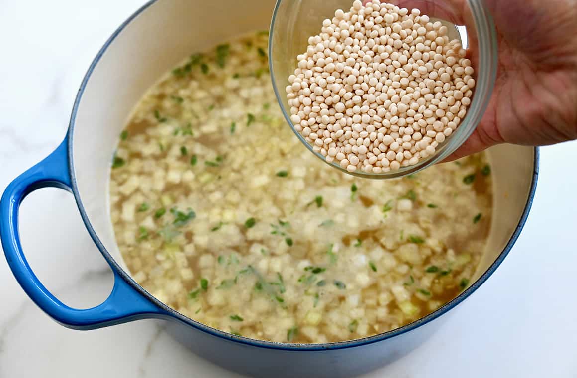 A hand holds a measuring cup filled with couscous over a large stockpot