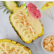 A top-down view of coconut pineapple smoothies in hollowed-out pineapple "bowls" topped with banana slices and toasted coconut.