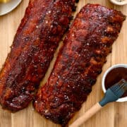 A top-down view of Oven-Baked Baby Back Ribs brushed with barbecue sauce next to a plate with cornbread and a bowl containing sliced pickles.