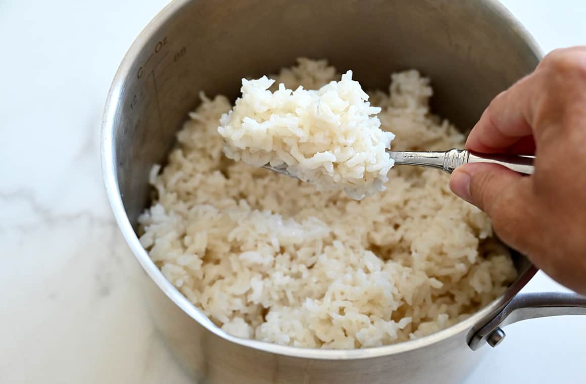 A hand holds a fork with cooked white rice over a pot containing cooked rice