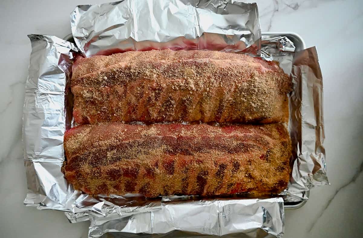 Unbaked baby back rib slabs with a dry spice rub atop a foil-lined baking sheet.