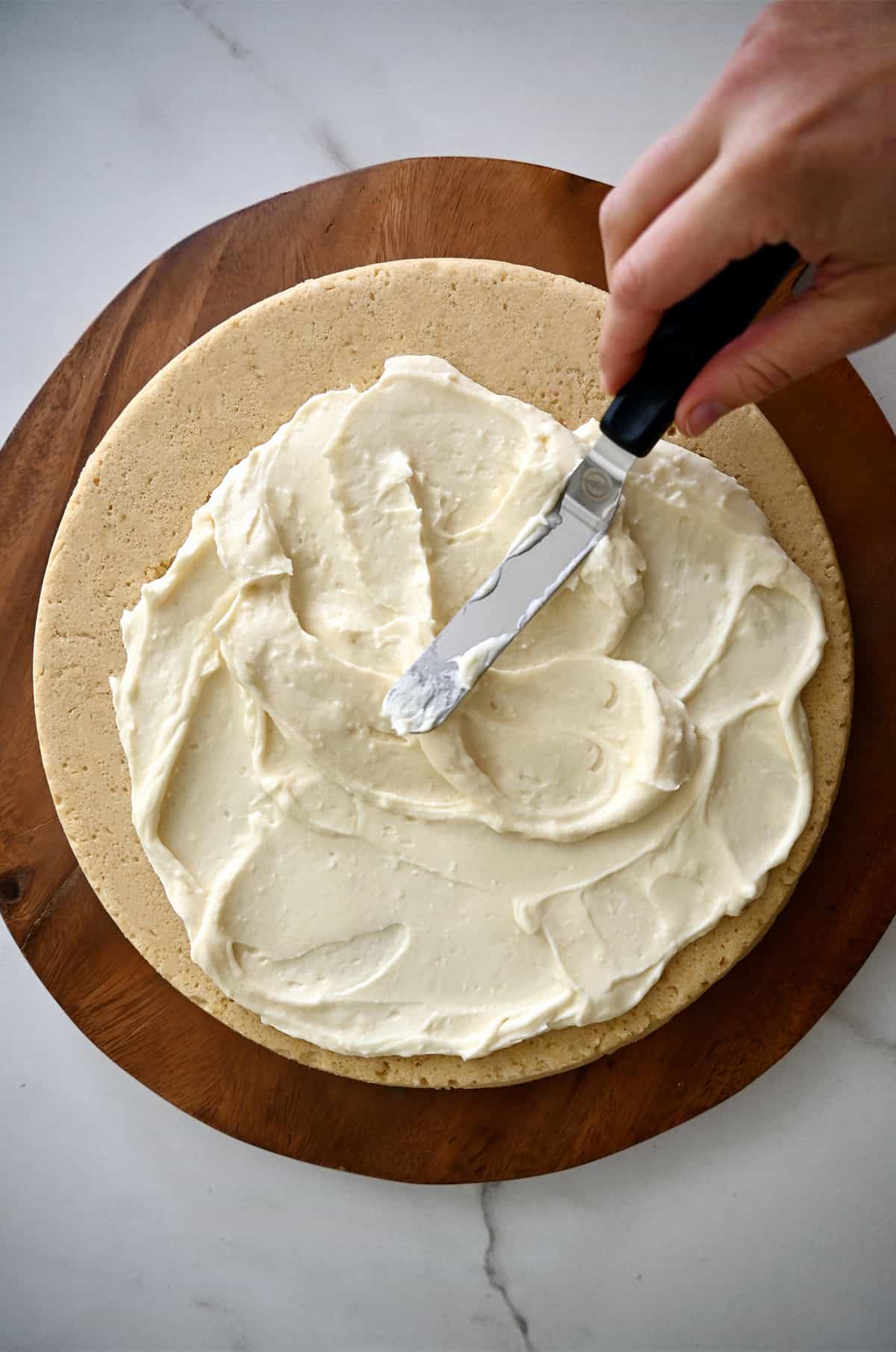 A hand holding an offset spatula spreads cream cheese frosting atop a giant sugar cookie.