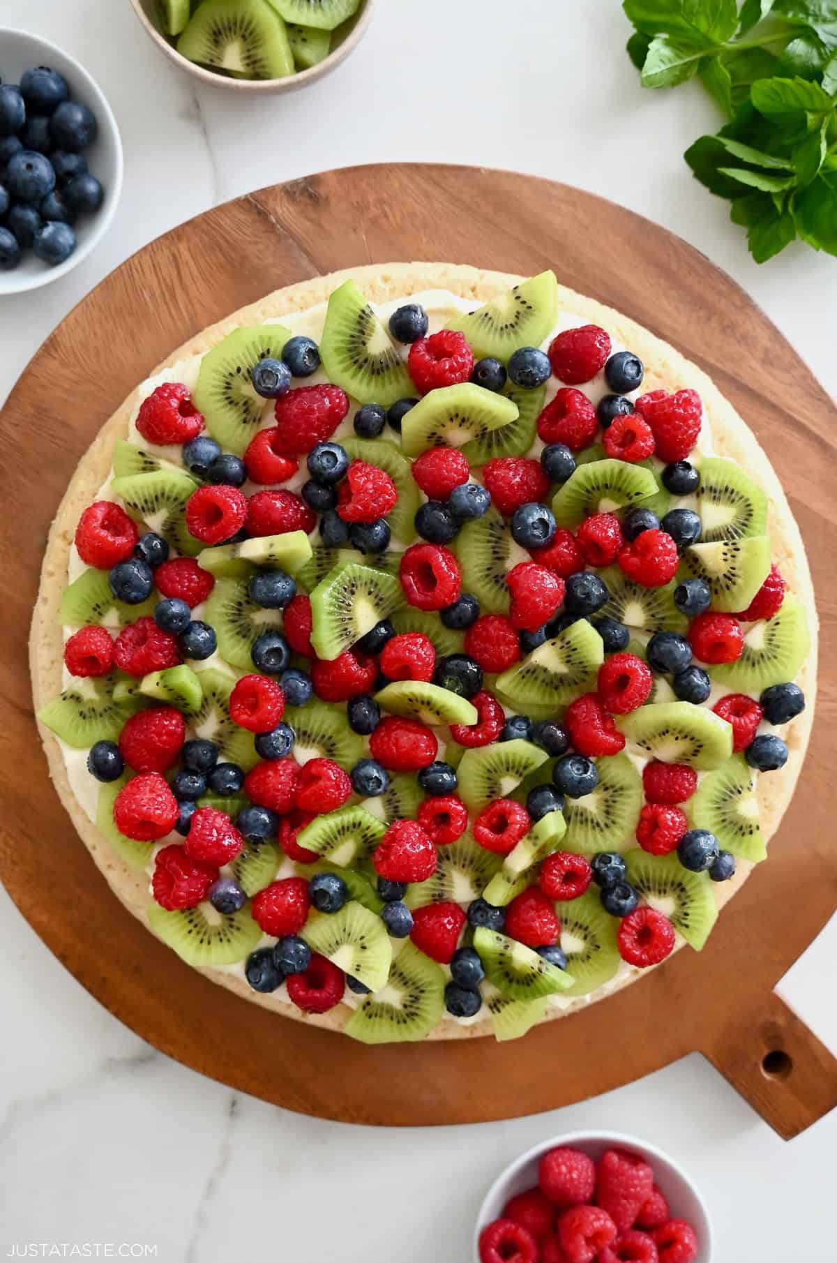 Sugar cookie fruit pizza with cream cheese frosting and fresh raspberries, blueberries and kiwi on a round platter.