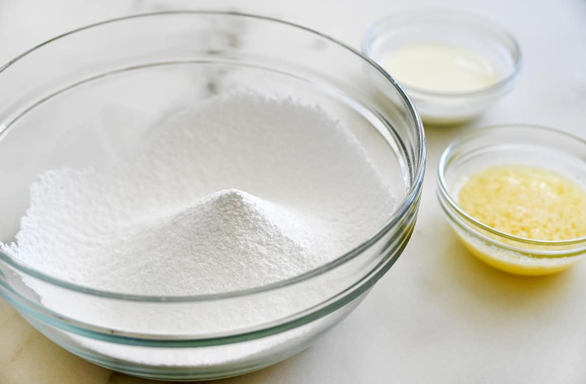 A clear bowl containing confectioners’ sugar next to two small bowls containing melted butter and whole milk