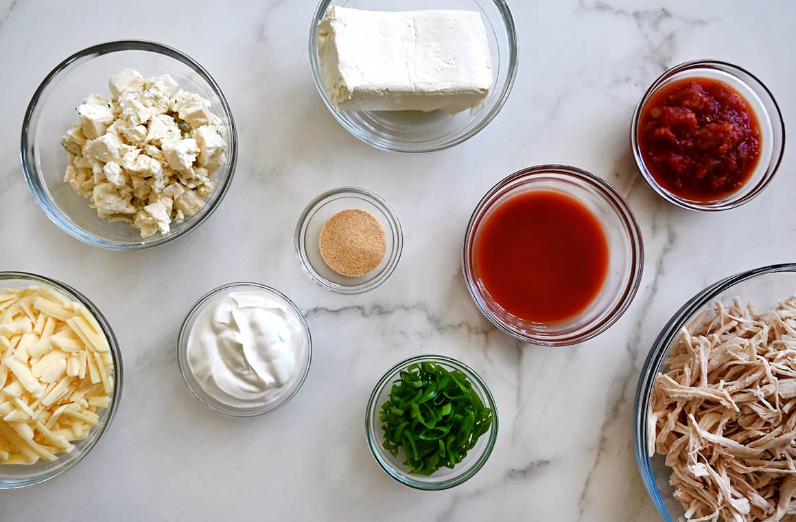A top-down view of various sizes of bowls containing cream cheese, hot sauce, shredded cooked chicken, sliced scallions, sour cream, garlic powder, blue cheese crumbles and shredded cheddar cheese