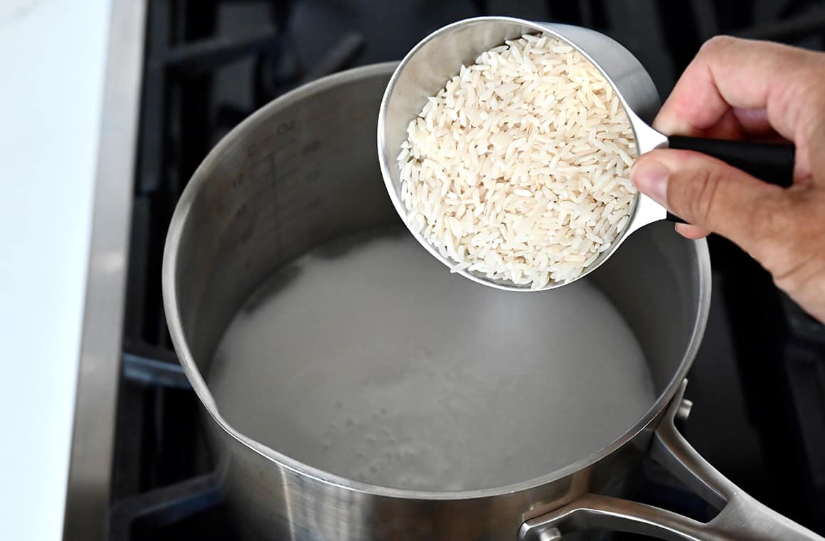 A hand holds a measuring cup filled with uncooked rice over a pot of water