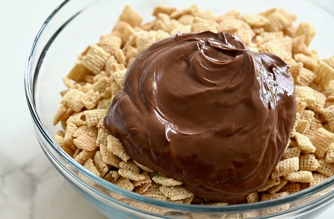 Melted chocolate chips and peanut butter over Chex Mix cereal in a clear bowl