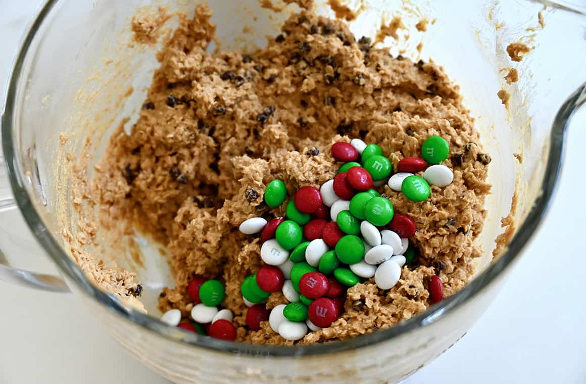 Red, white and green M&Ms atop chocolate chip dough in a clear bowl