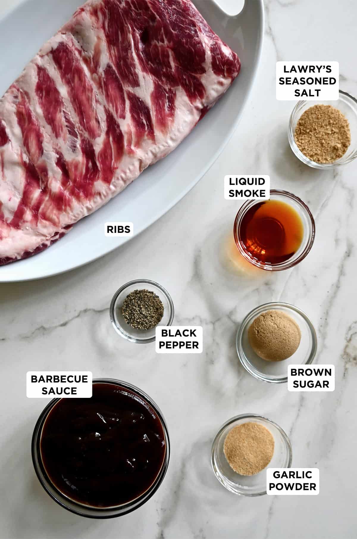 A rack of ribs on a plate next to small bowls containing barbecue sauce, black pepper, brown sugar, garlic powder, liquid smoke, and Lawry’s Seasoned Salt.