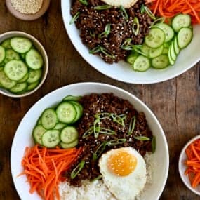 A top-down view of two bowls containing Korean beef, white rice, shredded carrots, sliced cucumber and a fried egg