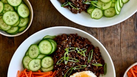 A top-down view of two bowls containing Korean beef, white rice, shredded carrots, sliced cucumber and a fried egg