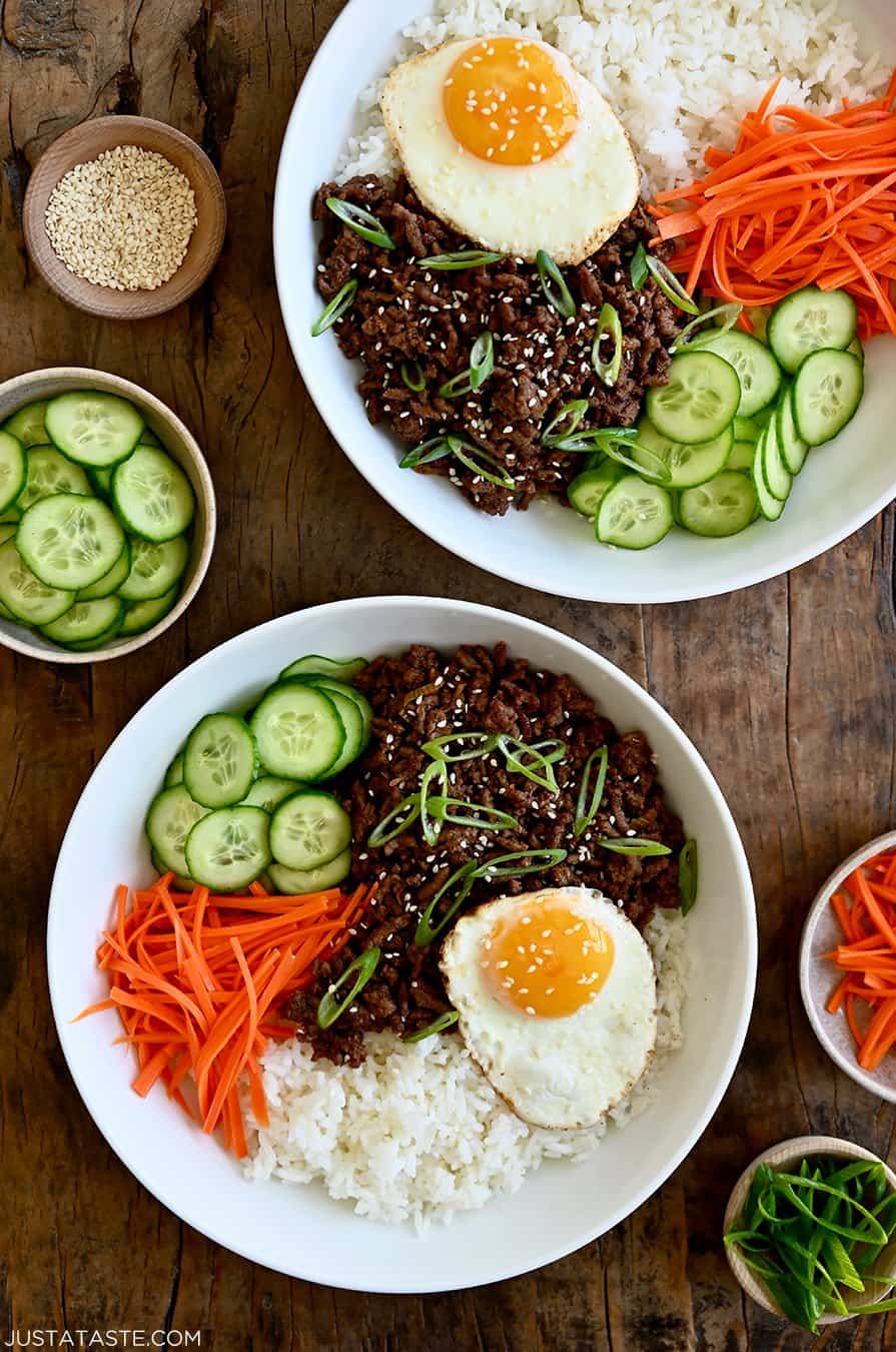 A top-down view of two bowls containing Korean beef, white rice, shredded carrots, sliced cucumber and a fried egg