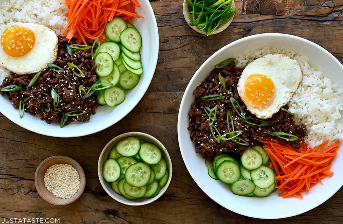 A top-down view of two white bowls containing quick Korean beef, jasmine rice, sliced carrots, sliced cucumber and a fried egg