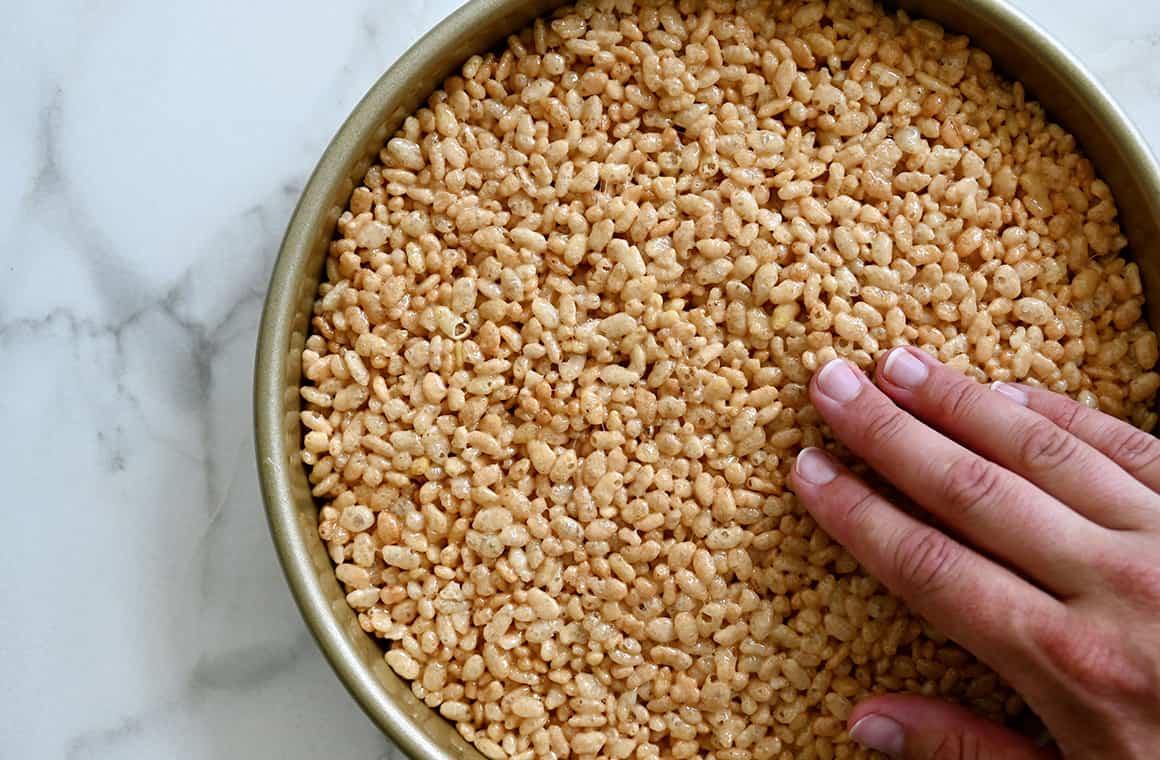 A hand presses marshmallow cereal mixture into a round cake pan