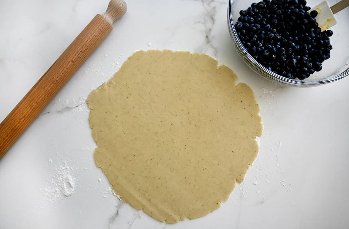 Rolled-out thyme pie dough next to a rolling pin and a glass bowl containing fresh blueberries.