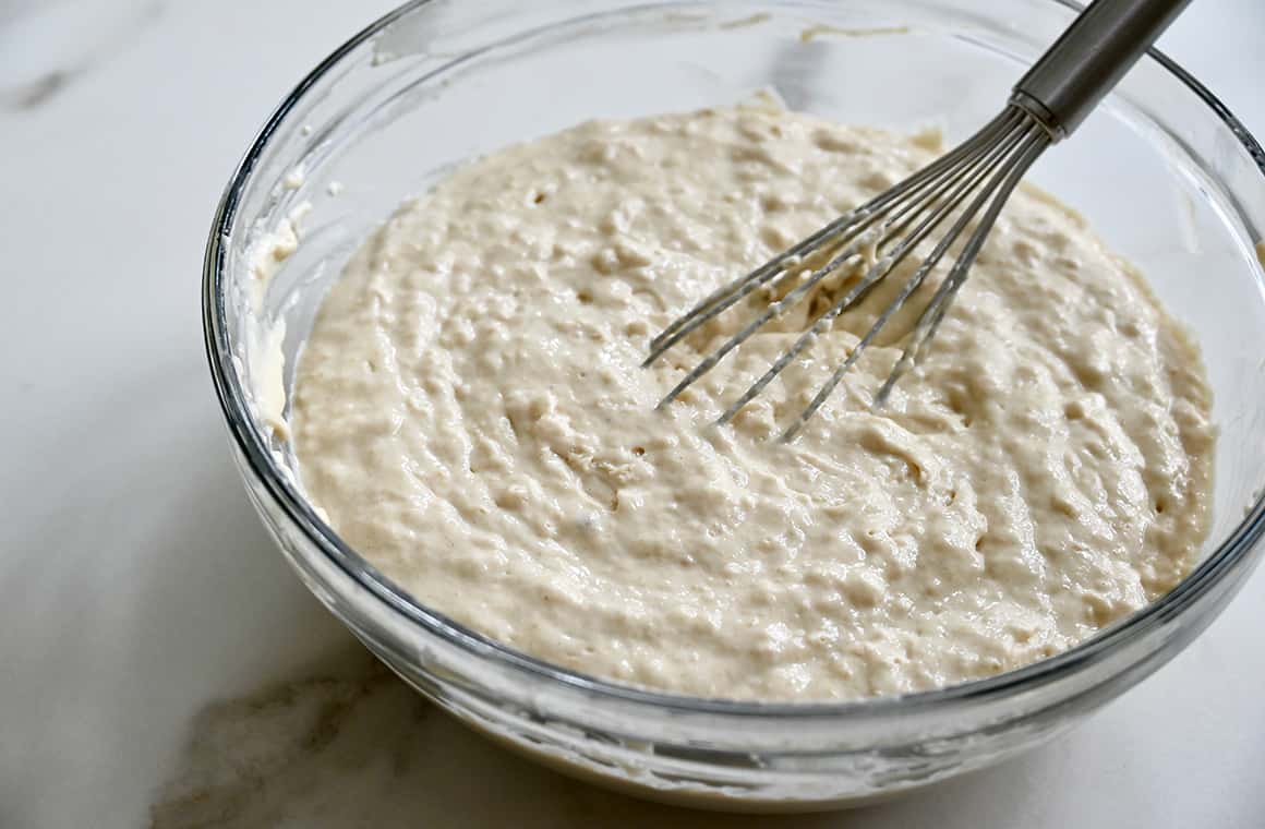 A whisk rests on the side of a clear bowl containing pancake batter