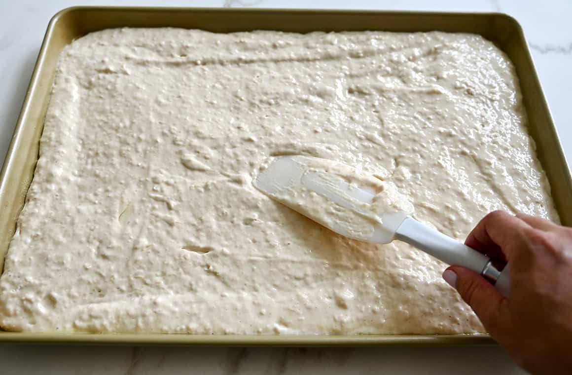 A hand holding a spatula spreads batter atop a baking sheet