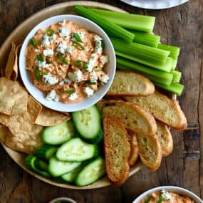 A top-down view of Slow Cooker Buffalo Chicken Dip in a small bowl on a plate surrounded by celery sticks, baguette toasts, sliced cucumber and tortilla chips
