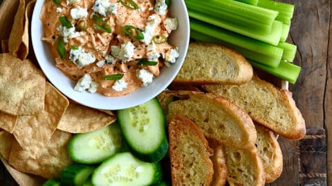 A top-down view of Slow Cooker Buffalo Chicken Dip in a small bowl on a plate surrounded by celery sticks, baguette toasts, sliced cucumber and tortilla chips