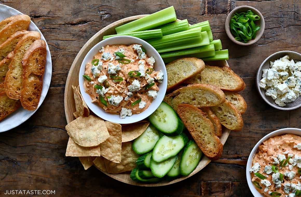 Slow Cooker Buffalo Chicken Dip garnished with blue cheese in a small bowl on a plate surrounded by baguette toasts, celery sticks, sliced cucumber and tortilla chips
