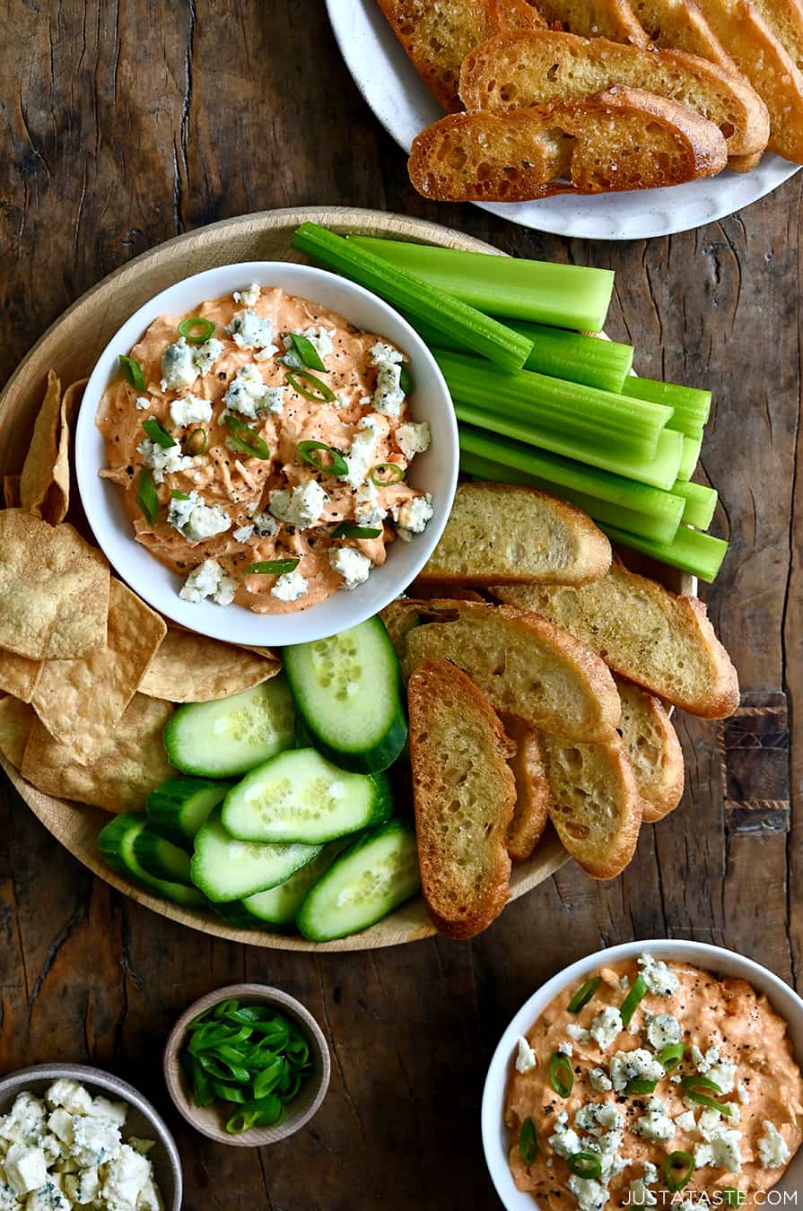 A top-down view of Slow Cooker Buffalo Chicken Dip in a small bowl on a plate surrounded by celery sticks, baguette toasts, sliced cucumber and tortilla chips