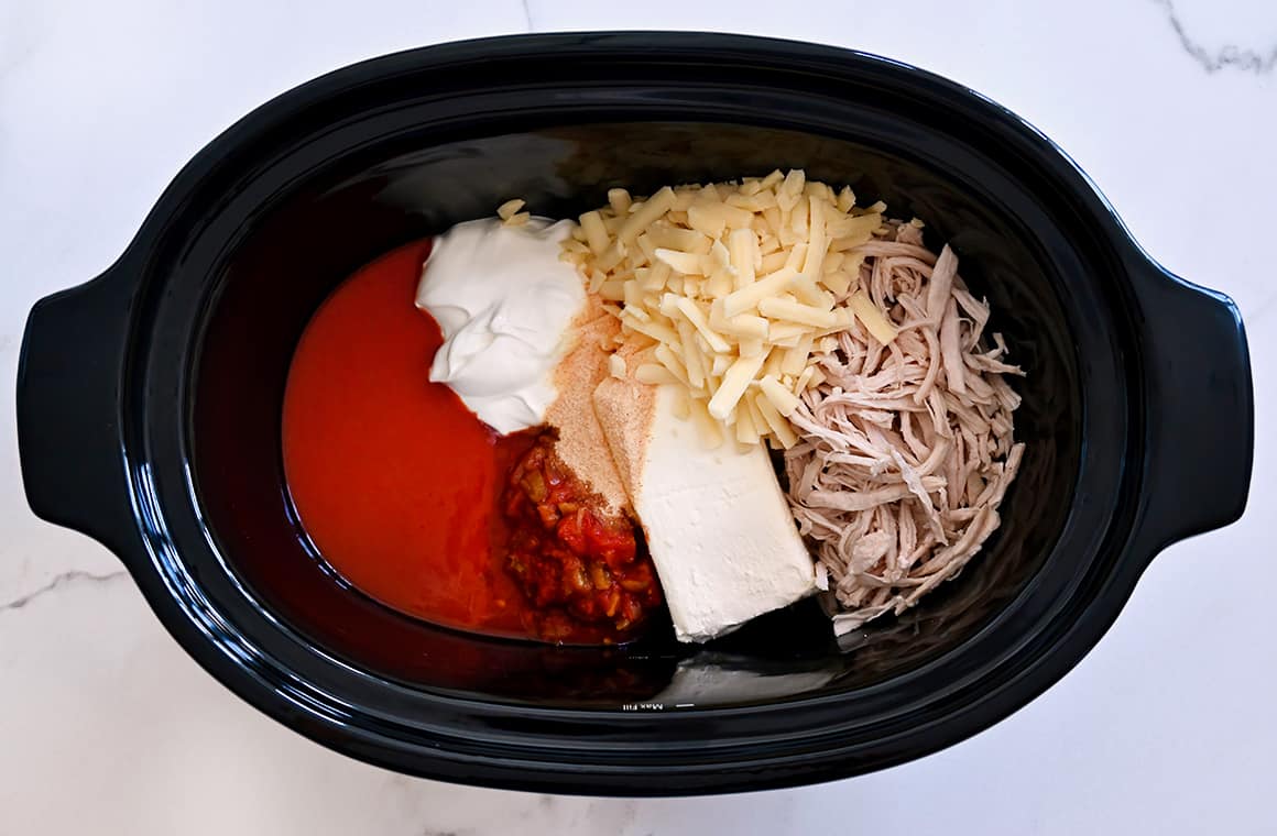 A top-down view of a slow cooker containing shredded cheddar cheese, shredded cooked chicken, a block of cream cheese, hot sauce and sour cream