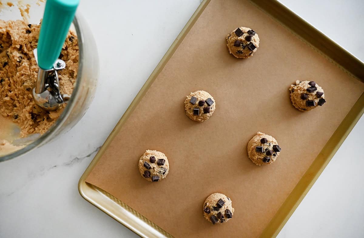 Six scoops of chocolate chip cookie dough on a parchment paper-lined baking sheet.