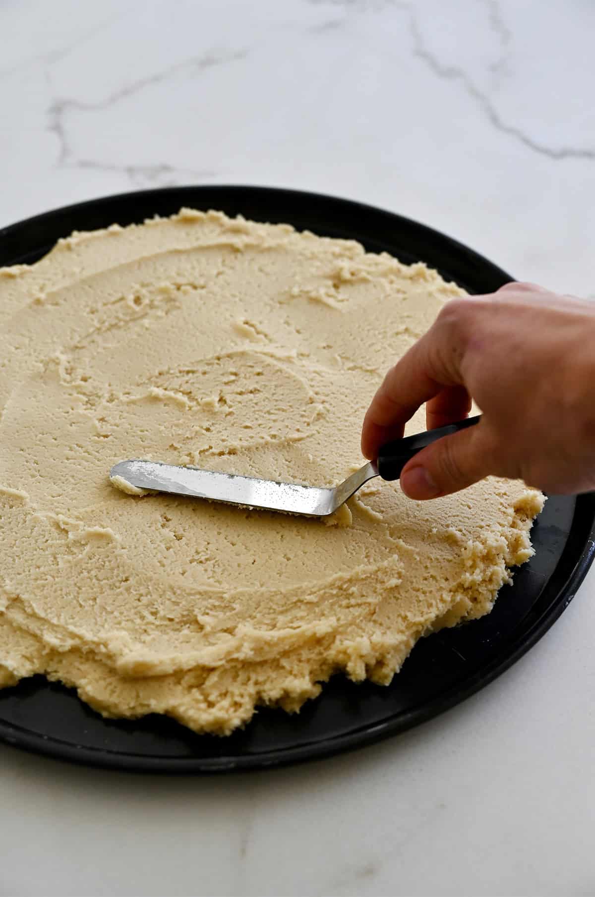 Using an offset spatula to spread sugar cookie dough into a circle on a pizza pan.