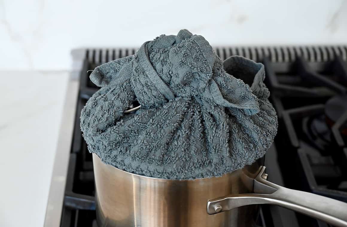 A medium pot covered with a towel-wrapped lid on a gas stovetop