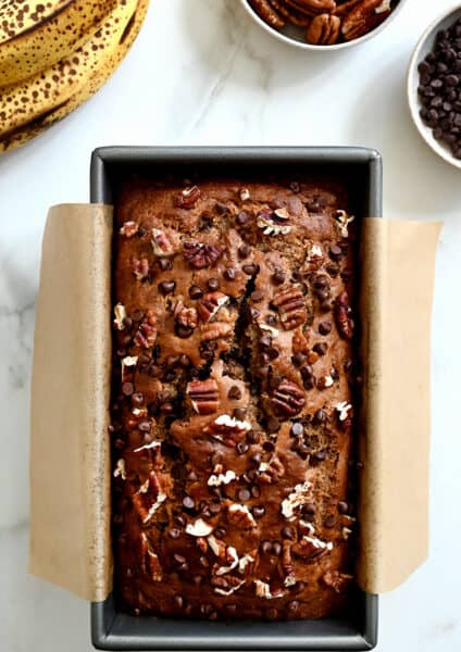 A top-down view of whole wheat banana bread topped with mini chocolate chips and chopped pecans in a parchment paper-lined bread pan