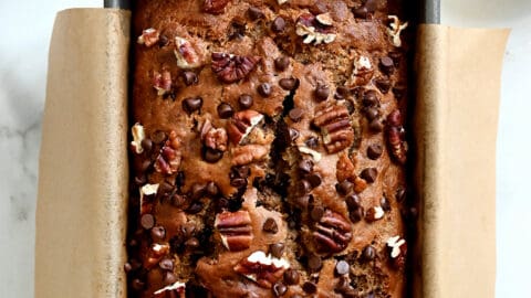 A top-down view of whole wheat banana bread topped with mini chocolate chips and chopped pecans in a parchment paper-lined bread pan