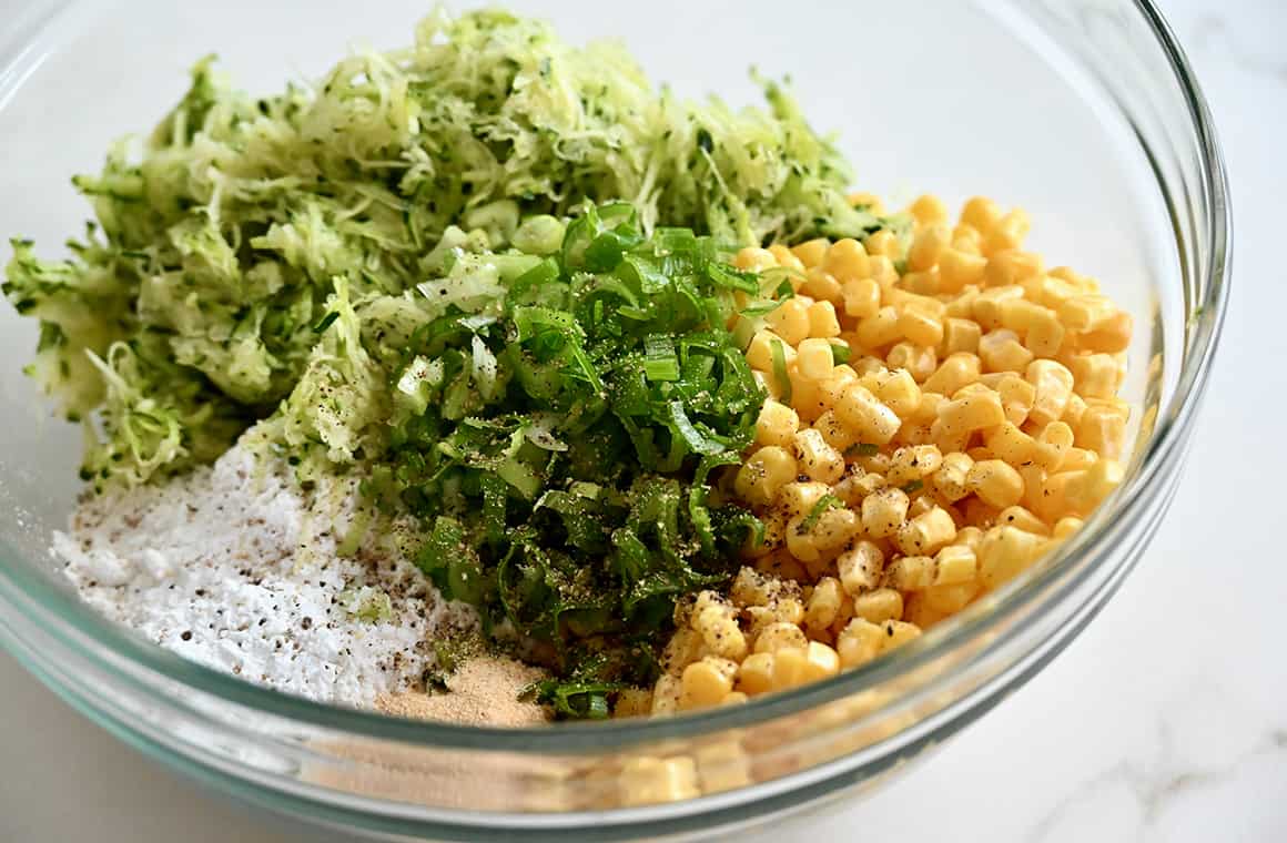 A clear bowl containing corn kernels, finely grated zucchini, chopped scallions and flour