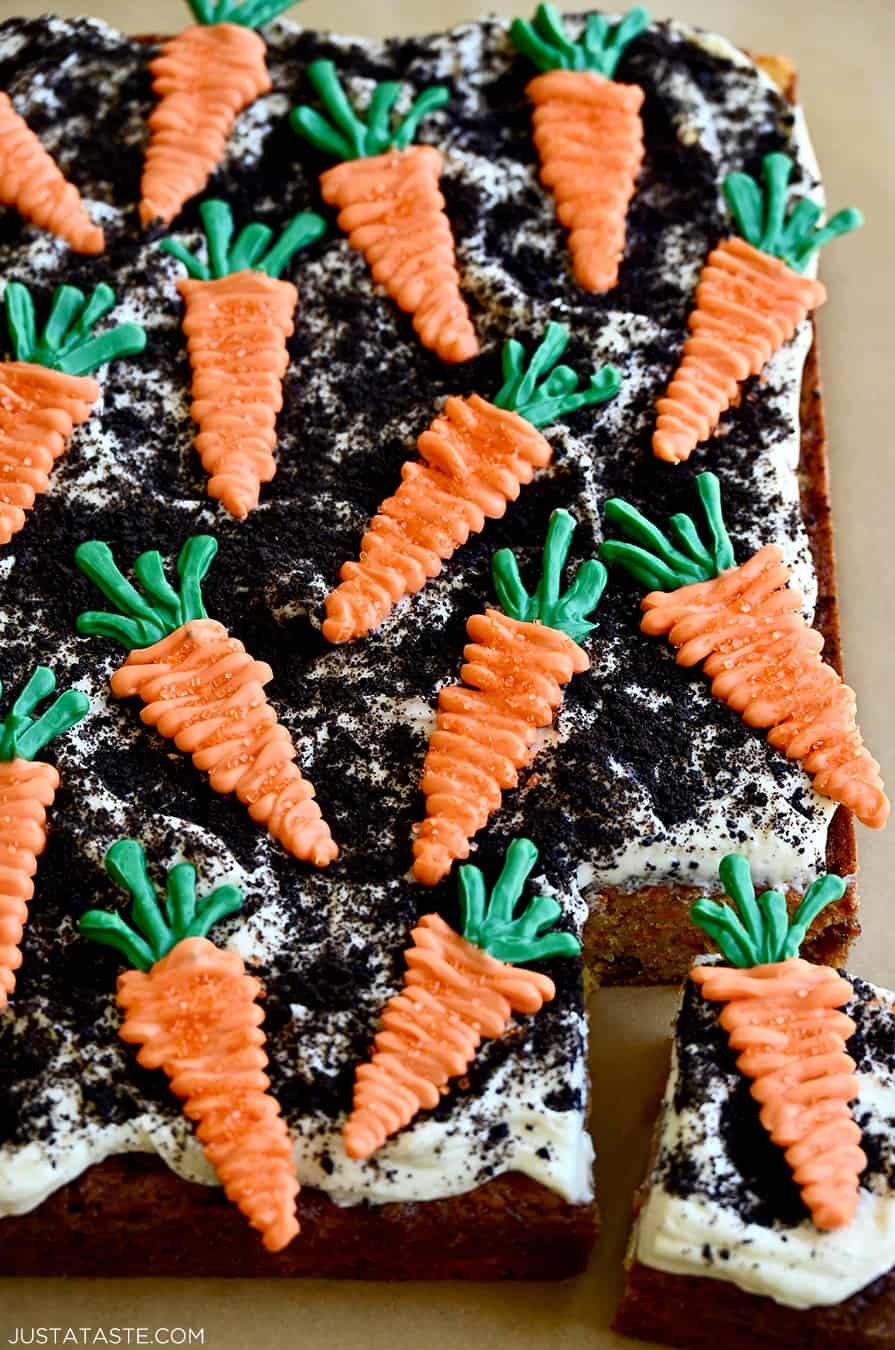 A corner slice of carrot sheet cake topped with cream cheese frosting, crushed Oreo "dirt" and candy melt "carrots"