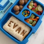 A top-down view of a Bentgo Kids lunchbox containing a peanut butter sandwich with the name "Evan," fruit, veggies, M&Ms and protein balls.