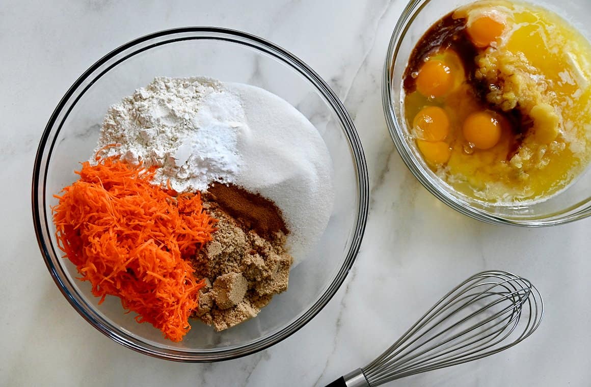 A top-down view of two clear bowls containing wet and dry ingredients, including flour, sugar, spices, eggs, oil and vanilla extract