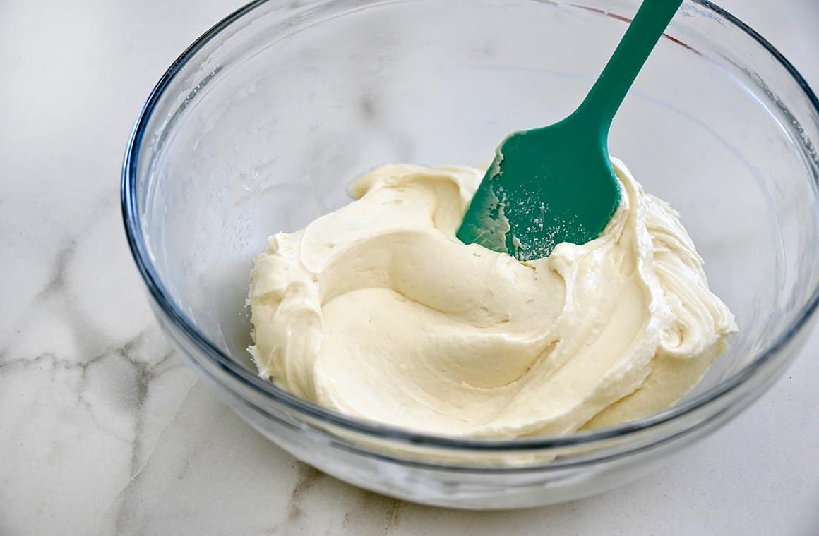 A clear bowl containing cream cheese frosting and a teal spatula