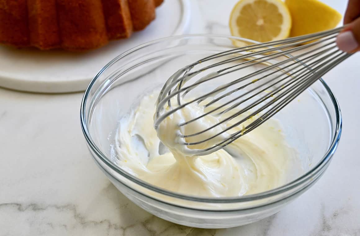 A clear bowl with icing and a wire whisk
