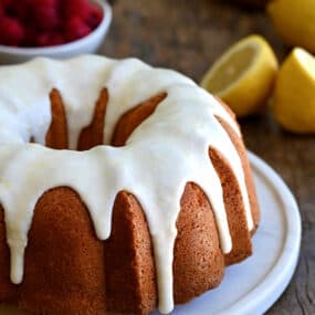 A glazed lemon pound cake on a round, white serving platter with a bowl filled with raspberries in the background