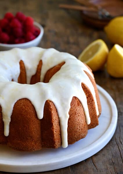 A glazed lemon pound cake on a round, white serving platter with a bowl filled with raspberries in the background