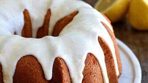 A glazed lemon pound cake on a round, white serving platter with a bowl filled with raspberries in the background