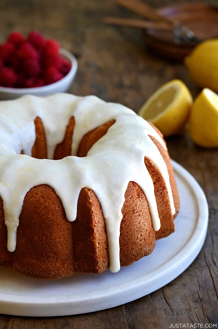 A glazed lemon pound cake on a round, white serving platter with a bowl filled with raspberries in the background