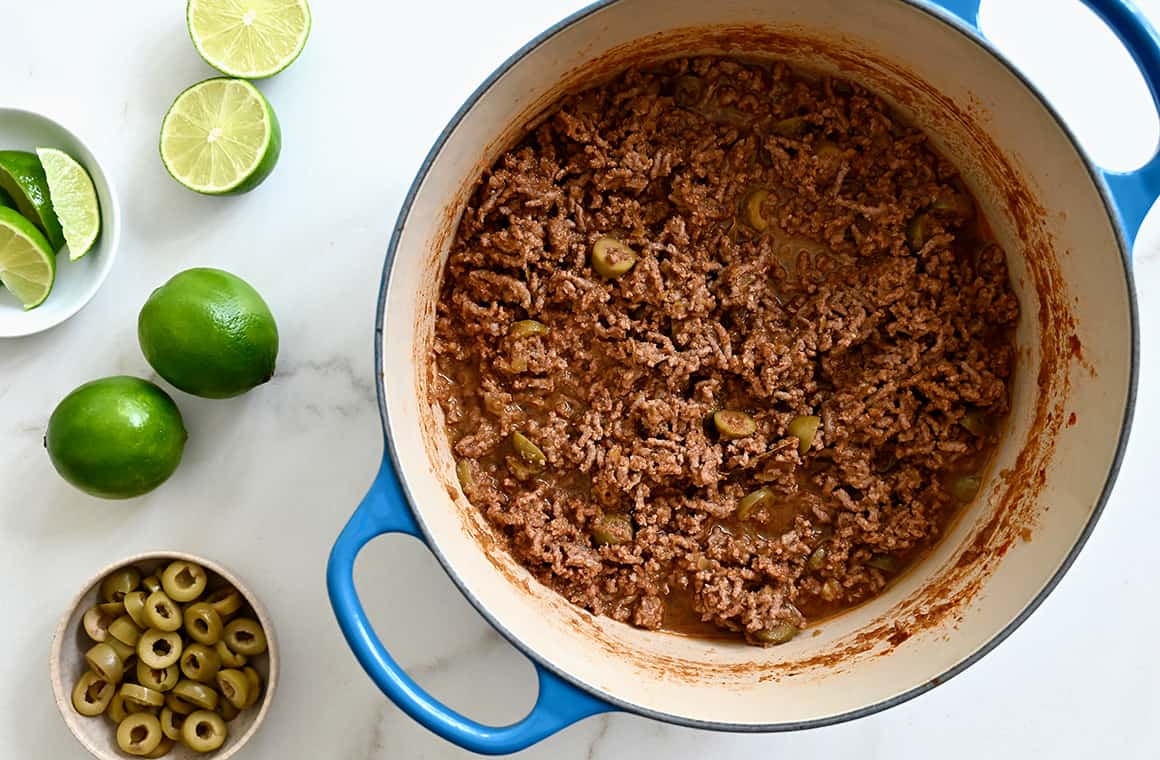 A top-down view of a large stockpot containing ground beef next to limes and a small bowl containing sliced green olives