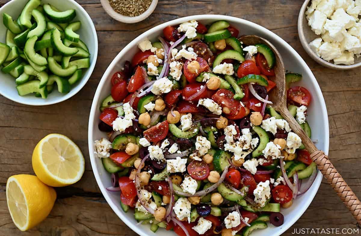 Cucumber, Tomato and Chickpea Salad with black olives and feta cheese in a white serving bowl with a wooden spoon.